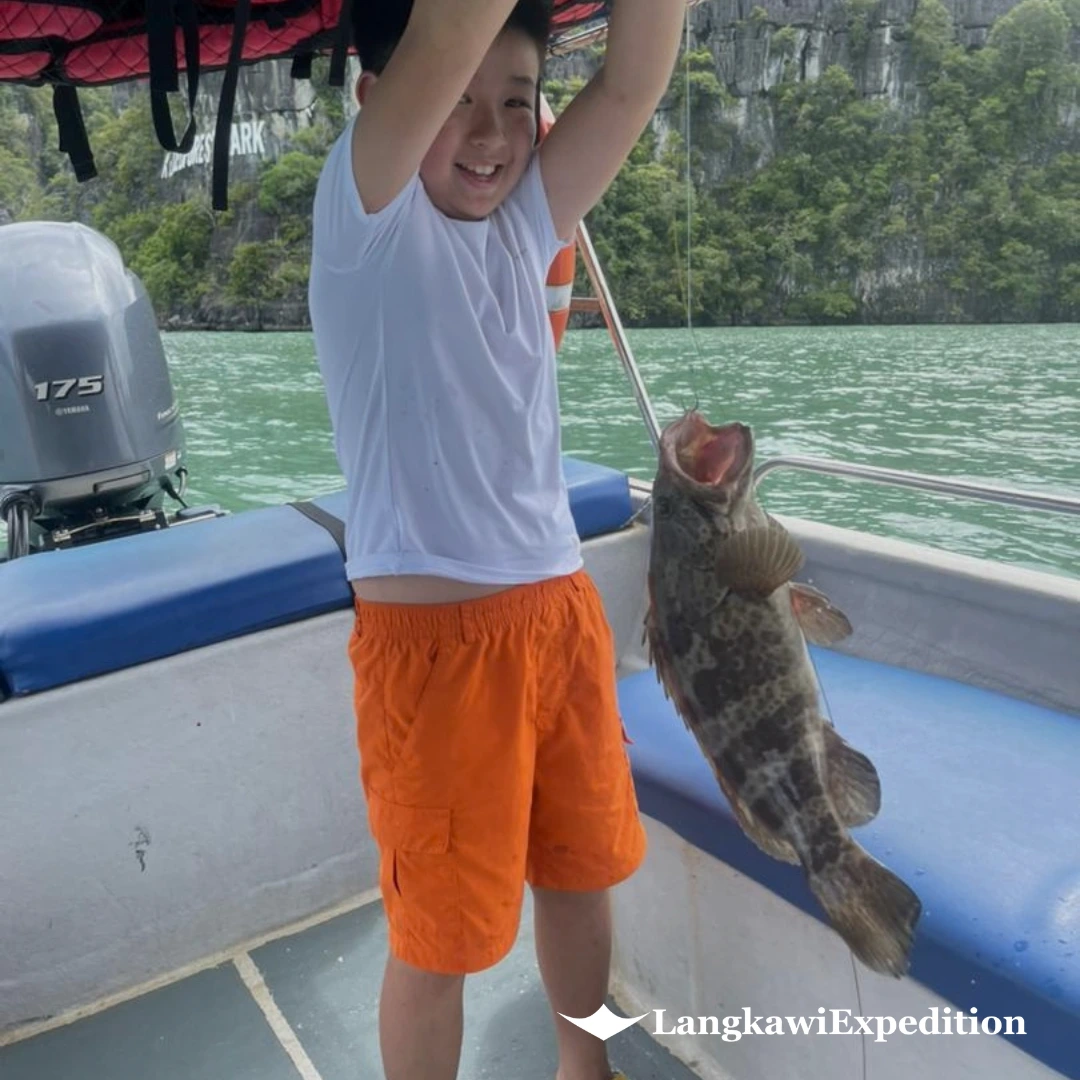 Young guest proudly holding a large grouper fish catch on a Langkawi fun fishing boat charter